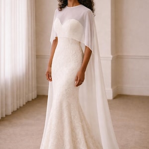 Beautiful bride in a modern white chiffon evening cape dress with a flowing sheer overlay and elegant satin base, standing gracefully against a soft beige background with warm studio lights highlighting her natural beauty. The chiffon cape adds ethereal movement, perfect for bridal photoshoots, rehearsal dinners, or wedding receptions. A minimalist yet luxurious fashion choice for the modern bride seeking timeless elegance.