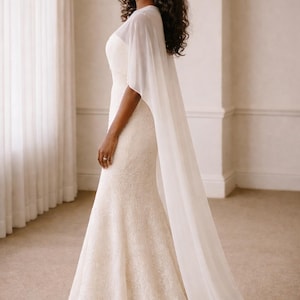 Beautiful bride in a modern white chiffon evening cape dress with a flowing sheer overlay and elegant satin base, standing gracefully against a soft beige background with warm studio lights highlighting her natural beauty. The chiffon cape adds ethereal movement, perfect for bridal photoshoots, rehearsal dinners, or wedding receptions. A minimalist yet luxurious fashion choice for the modern bride seeking timeless elegance.