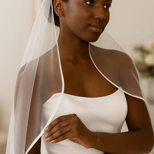 Close-up image of a dark-skinned bridal model smiling warmly while wearing a satin edge veil. The soft beige background highlights her radiant expression and the elegant detailing of the veil.