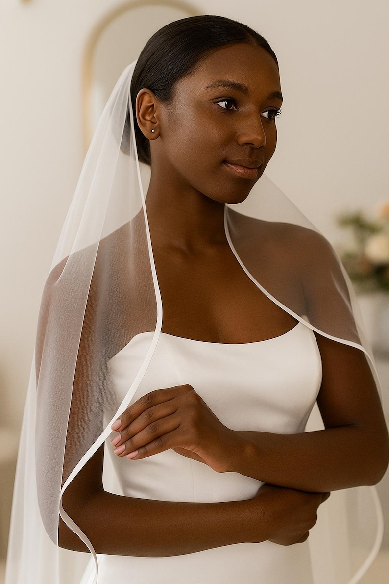 Close-up image of a dark-skinned bridal model smiling warmly while wearing a satin edge veil. The soft beige background highlights her radiant expression and the elegant detailing of the veil.