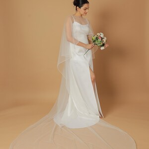 A bride wearing an elegant lace wedding gown is adorned with a sheer, white tulle bridal cape. The cape features a soft round neckline and flows gracefully over her shoulders, ending just above the waist in front and trailing longer in the back. The fitted gown has a subtle floral texture and a strapless sweetheart neckline. The setting is softly lit with neutral tones, featuring white curtains and minimal décor, highlighting the romantic and refined look.