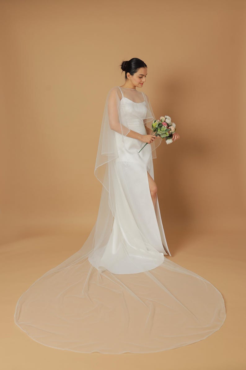 A bride wearing an elegant lace wedding gown is adorned with a sheer, white tulle bridal cape. The cape features a soft round neckline and flows gracefully over her shoulders, ending just above the waist in front and trailing longer in the back. The fitted gown has a subtle floral texture and a strapless sweetheart neckline. The setting is softly lit with neutral tones, featuring white curtains and minimal décor, highlighting the romantic and refined look.