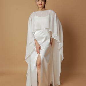 May include: A white satin gown with a high slit, complemented by a sheer cape. The dress has a simple, elegant design, suitable for formal occasions. The model is wearing clear sandals.