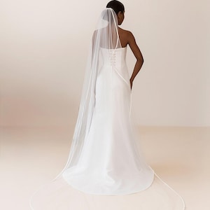 Backside image of a dark-skinned bridal model while wearing a Ivory tulle satin edge veil. The soft beige background highlights her radiant expression and the elegant detailing of the veil.