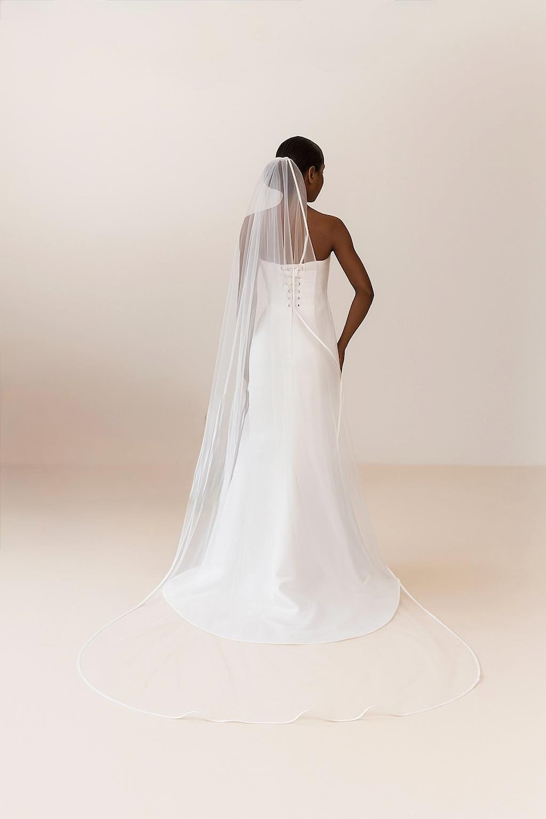 Backside image of a dark-skinned bridal model while wearing a Ivory tulle satin edge veil. The soft beige background highlights her radiant expression and the elegant detailing of the veil.