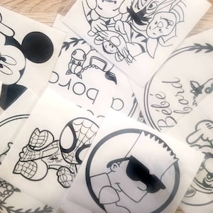 May include: A collection of white stickers featuring black cartoon character designs. The stickers include Mickey Mouse, Minnie Mouse, Spider-Man, Bart Simpson, and other cartoon characters. Some stickers have text, including "Bébé à bord".