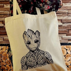 Könnte beinhalten: Cremefarbene Tragetasche mit einer schwarzen Grafik einer Groot-ähnlichen Figur. Das Design umfasst detaillierte Gesichtszüge und Muster im Tribal-Stil auf Brust und Schultern. Die Tasche hat lange, cremefarbene Träger.