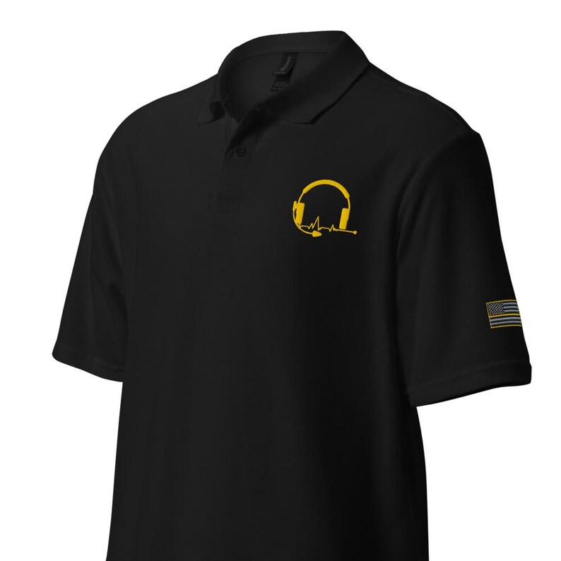 May include: Black polo shirt with a yellow embroidered headset and a heartbeat graphic. The shirt also features a thin yellow American flag on the right sleeve.