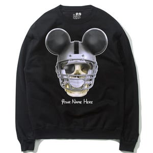 Puede incluir: Sudadera negra con una calavera blanca que lleva un casco de f&uacute;tbol americano y orejas de Mickey Mouse. El texto "Your Name Here" est&aacute; impreso debajo de la calavera.