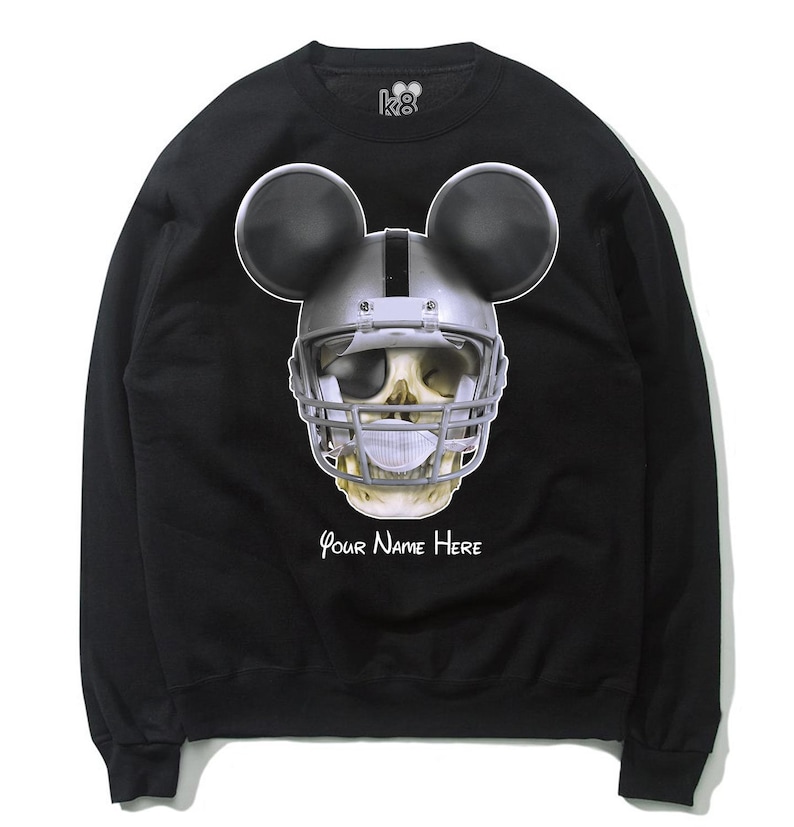 Puede incluir: Sudadera negra con una calavera blanca que lleva un casco de f&uacute;tbol americano y orejas de Mickey Mouse. El texto "Your Name Here" est&aacute; impreso debajo de la calavera.