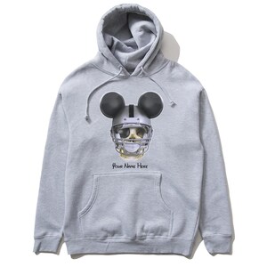 Puede incluir: Una sudadera con capucha gris con una calavera en blanco y negro que lleva un casco de f&uacute;tbol americano y orejas de Mickey Mouse. El texto "Your Name Here" est&aacute; impreso debajo de la calavera.