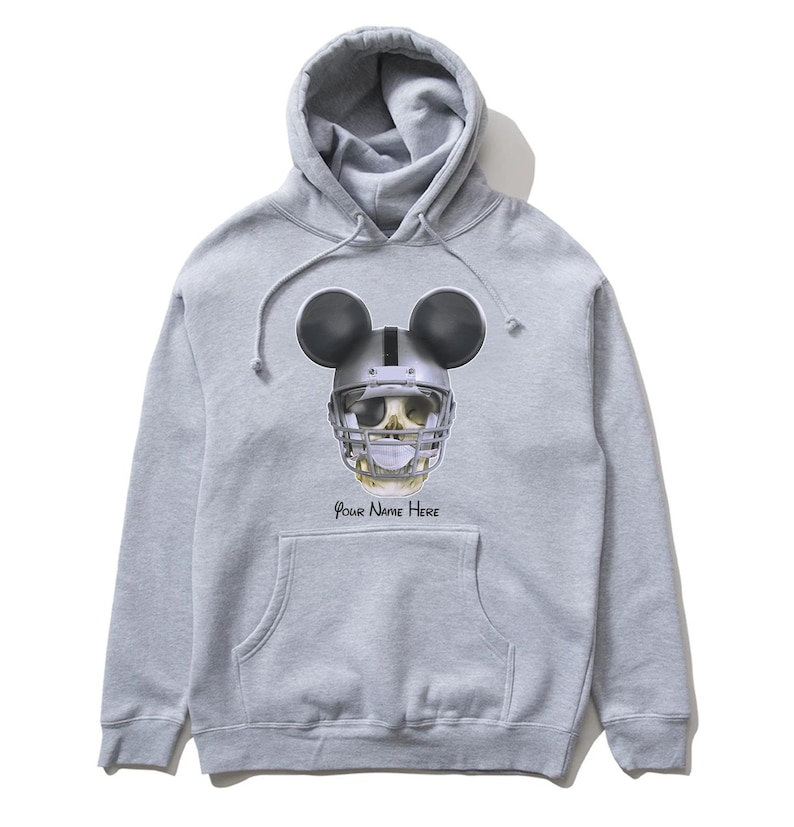Puede incluir: Una sudadera con capucha gris con una calavera en blanco y negro que lleva un casco de f&uacute;tbol americano y orejas de Mickey Mouse. El texto "Your Name Here" est&aacute; impreso debajo de la calavera.