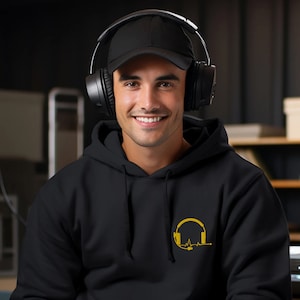 May include: A black hoodie with a gold headphone and heartbeat graphic. The person wearing the hoodie is smiling and wearing a black baseball cap and black headphones.