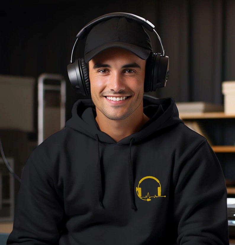 May include: A black hoodie with a gold headphone and heartbeat graphic. The person wearing the hoodie is smiling and wearing a black baseball cap and black headphones.