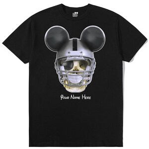 Puede incluir: Camiseta negra con un gr&aacute;fico de una calavera con un casco de f&uacute;tbol americano y orejas de Mickey Mouse. El texto "Your Name Here" est&aacute; impreso debajo del gr&aacute;fico.