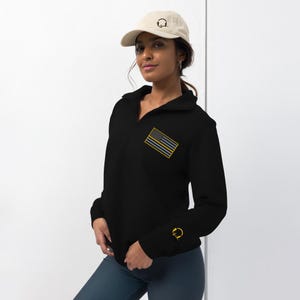 May include: Black mock neck sweatshirt with a thin yellow stripe across the American flag on the left chest. The sweatshirt has a zipper at the neck and a small gold logo on the right sleeve.