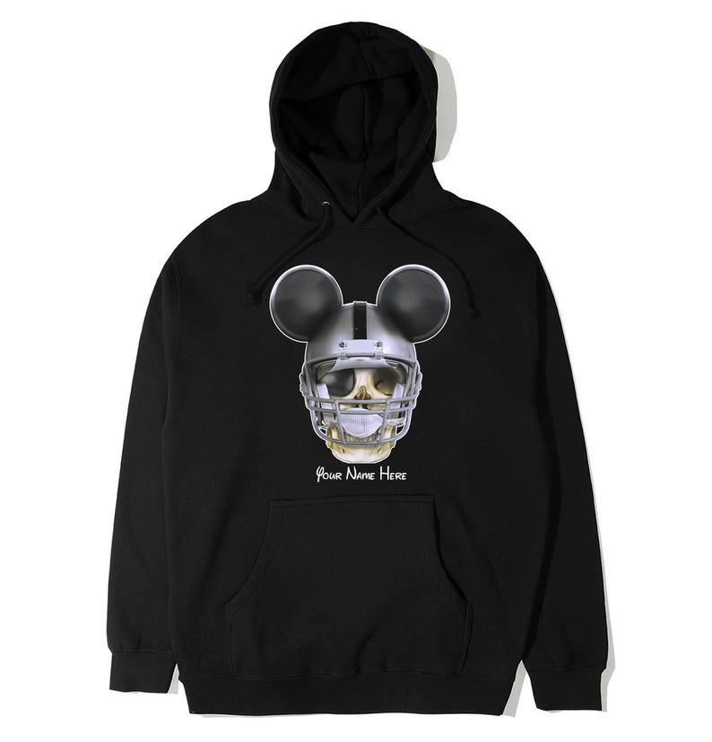 Puede incluir: Sudadera con capucha negra con una calavera con un casco de f&uacute;tbol americano y orejas de Mickey Mouse. El texto "Your Name Here" est&aacute; impreso debajo de la calavera.