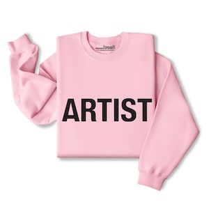 Puede incluir: Una sudadera rosa con la palabra "ARTIST" impresa en negro en la parte delantera.