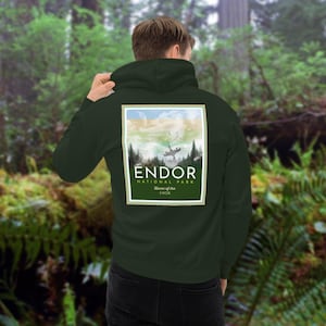 Puede incluir: Una sudadera con capucha verde oscuro con un diseño de póster de parque nacional de estilo vintage en la espalda. El póster presenta una ilustración de acuarela de una escena de bosque con una nave espacial volando sobre un cielo nublado. El texto en el póster dice "Endor National Park, Home of the Ewok".