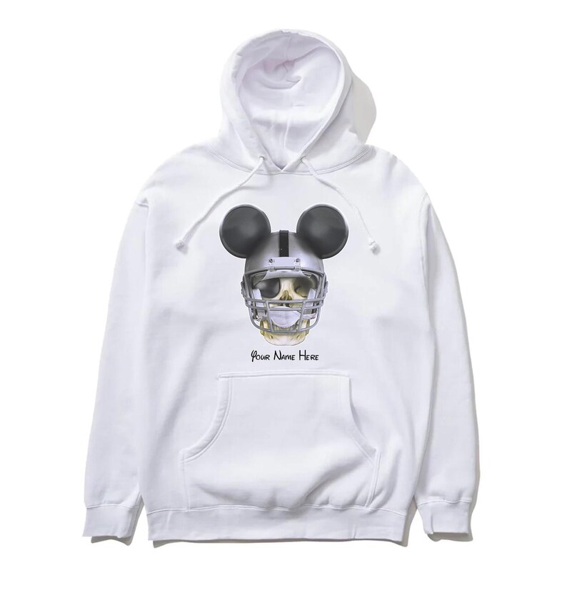 Puede incluir: Sudadera con capucha blanca con una calavera en blanco y negro que lleva un casco de f&uacute;tbol americano y orejas de Mickey Mouse. El texto "Your Name Here" est&aacute; impreso debajo de la calavera.