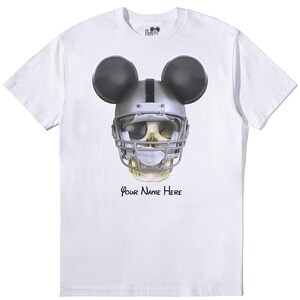 Puede incluir: Camiseta blanca con un gr&aacute;fico de una calavera con un casco de f&uacute;tbol americano y orejas de Mickey Mouse. El texto "Your Name Here" est&aacute; impreso debajo del gr&aacute;fico.