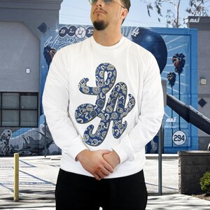 May include: White long-sleeve sweatshirt with a large, stylized "LA" graphic in blue paisley print. The sweatshirt is worn by a person wearing sunglasses. The background features a mural with palm trees and the number 294.