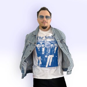 May include: A white t-shirt with a blue print of the band "The Smiths" on the front. The person wearing the shirt is also wearing a light blue denim jacket.