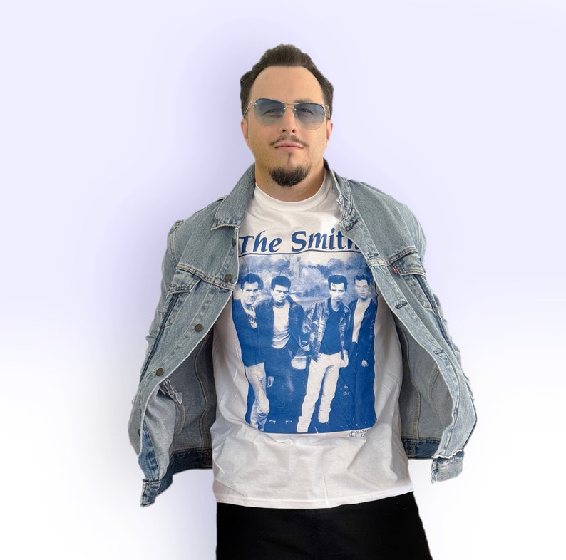 May include: A white t-shirt with a blue print of the band "The Smiths" on the front. The person wearing the shirt is also wearing a light blue denim jacket.