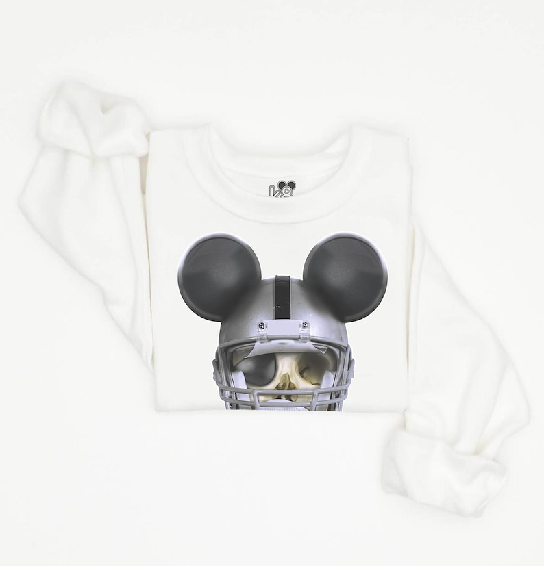 Puede incluir: Camiseta blanca de manga larga con una calavera negra y plateada que lleva un casco de f&uacute;tbol americano y orejas de Mickey Mouse.