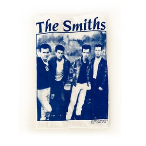 May include: A white t-shirt with a blue print of the band The Smiths. The band members are pictured in black and white, standing in front of a building. The text "The Smiths" is printed in large blue letters at the top of the shirt. The text "Manchester England" is printed in smaller blue letters at the bottom of the shirt.