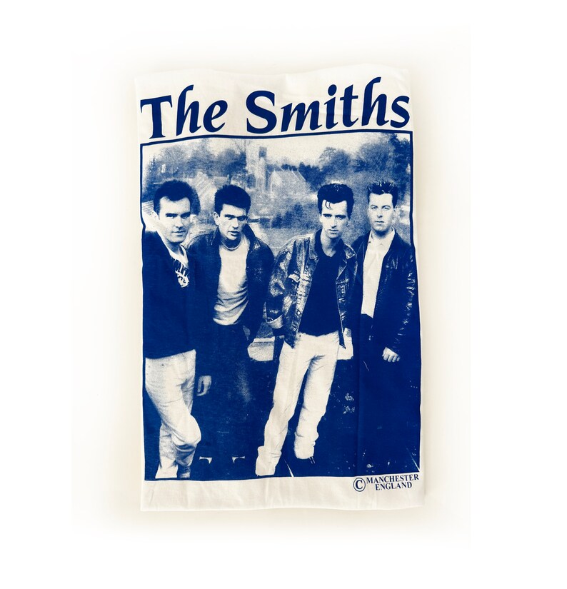 May include: A white t-shirt with a blue print of the band The Smiths. The band members are pictured in black and white, standing in front of a building. The text "The Smiths" is printed in large blue letters at the top of the shirt. The text "Manchester England" is printed in smaller blue letters at the bottom of the shirt.