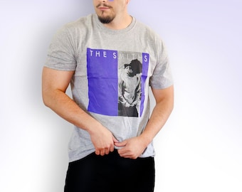 Può includere: Una maglietta grigia con un'immagine viola e bianca di una persona e il testo "THE SMITHS".