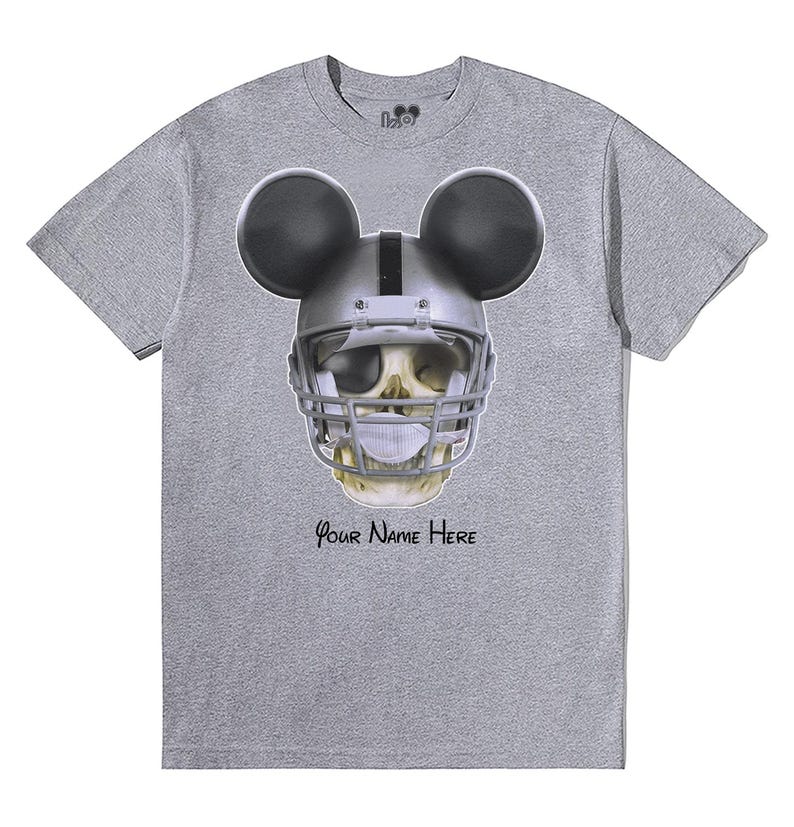 Puede incluir: Camiseta gris con una calavera con un casco de f&uacute;tbol americano y orejas de Mickey Mouse. El texto "Your Name Here" est&aacute; impreso debajo de la calavera.