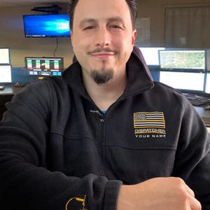 May include: Black zip-up fleece jacket with a gold and black American flag patch that says "DISPATCHER YOUR NAME". The jacket also has a gold headphone graphic on the sleeve.