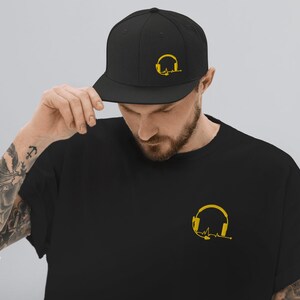 May include: A black t-shirt featuring a yellow graphic of headphones and a heartbeat. The t-shirt is worn with a black baseball cap with the same design.