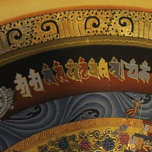 May include: A traditional Tibetan mandala painting with intricate details and a gold border. The painting features a central wheel of dharma, surrounded by various symbols and figures. The text at the top reads "Om Ah Hum."