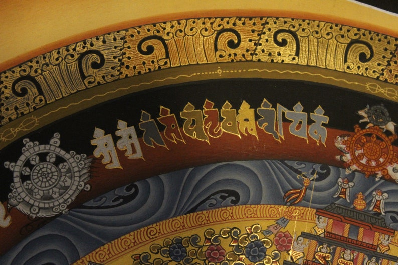 May include: A traditional Tibetan mandala painting with intricate details and a gold border. The painting features a central wheel of dharma, surrounded by various symbols and figures. The text at the top reads "Om Ah Hum."