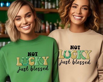 May include: Two women wearing sweatshirts with the text "NOT LUCKY just blessed". One sweatshirt is green, the other is tan. The text includes a horseshoe and shamrock design.