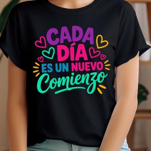 May include: Black t-shirt with the text "CADA DÍA ES UN NUEVO COMIENZO" in vibrant colors, including purple, pink, and teal. The design features heart shapes and sunburst accents, creating a cheerful and positive vibe.