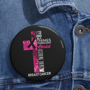 Puede incluir: Un botón redondo negro con una cruz rosa y un lazo negro de concienciación. El texto dice: I CAN DO ALL THINGS THROUGH Christ WHO STRENGTHENS ME BREAST CANCER. El botón está sujeto a una tela de mezclilla azul.