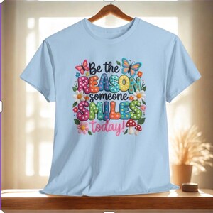 May include: A pale blue t-shirt featuring the phrase "Be the REASON someone SMILES today!" in a vibrant, multi-coloured design. The graphic includes butterflies, flowers, and mushrooms, giving a cheerful and positive feel.