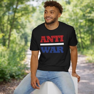Op de afbeelding: Zwart t-shirt met de woorden "ANTI WAR" in rode en blauwe blokletters, gescheiden door een witte horizontale lijn. De persoon draagt een spijkerbroek en zit op een wit oppervlak.
