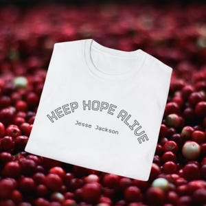 May include: White t-shirt featuring the text "KEEP HOPE ALIVE" and "Jesse Jackson" in black. The shirt is set against a backdrop of red berries.