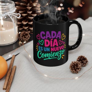 May include: A black ceramic mug with the Spanish phrase "Cada Día Es Un Nuevo Comienzo" in purple, pink, teal, and yellow. The mug has a curved handle and is filled with a dark liquid, possibly coffee or tea. The mug is on a light gray surface.