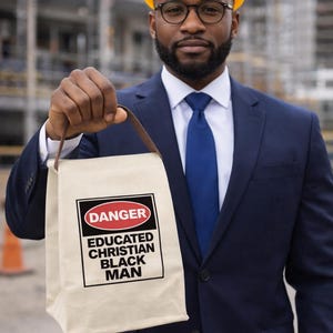 May include: A man in a navy blue suit, blue tie, and yellow hard hat holds a beige tote bag. The bag has a black and red danger sign with the words "EDUCATED CHRISTIAN BLACK MAN" in black text.