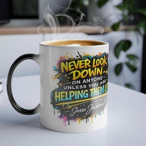May include: White ceramic mug with a black handle, filled with coffee and steam. The mug features the quote "Never Look Down On Anyone - Unless You Are Helping Them Up" in colorful graffiti-style lettering, signed by Jesse Jackson.