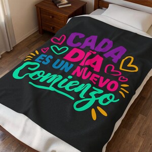 May include: Black blanket with the text "Cada Día Es Un Nuevo Comienzo" in vibrant colors. The design includes purple, pink, green, and yellow lettering with heart accents. The blanket is on a bed with a wooden nightstand.