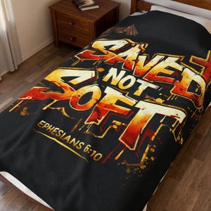 May include: Black blanket featuring the text "SAVED NOT SOFT" in a graffiti style, with orange and yellow hues. The words "EPHESIANS 6:10" are printed below. The blanket is displayed on a bed.