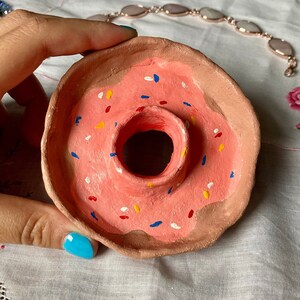 May include: A hand-painted ceramic donut-shaped trinket dish. The dish features a pink frosting with colorful sprinkles and a brown base. The center has a hole, and a bite is missing from the side. A unique and playful decorative item.