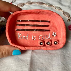 Op de afbeelding: Een handgemaakte, koraalkleurige kleien sieradenschaal. De schaal heeft drie donkerbruine strepen met witte accenten, de tekst "Kind is Cool", een kleine bloem en drie kleine, ronde versieringen. Sieraden zijn zichtbaar op de achtergrond.